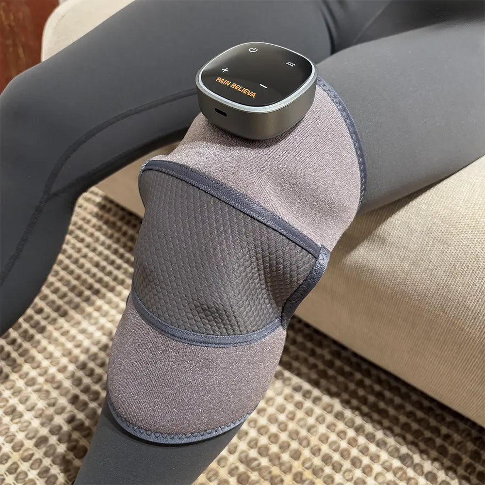 The PainRelieva™ Heated Knee Massager worn on a person's knee, sitting on a couch.